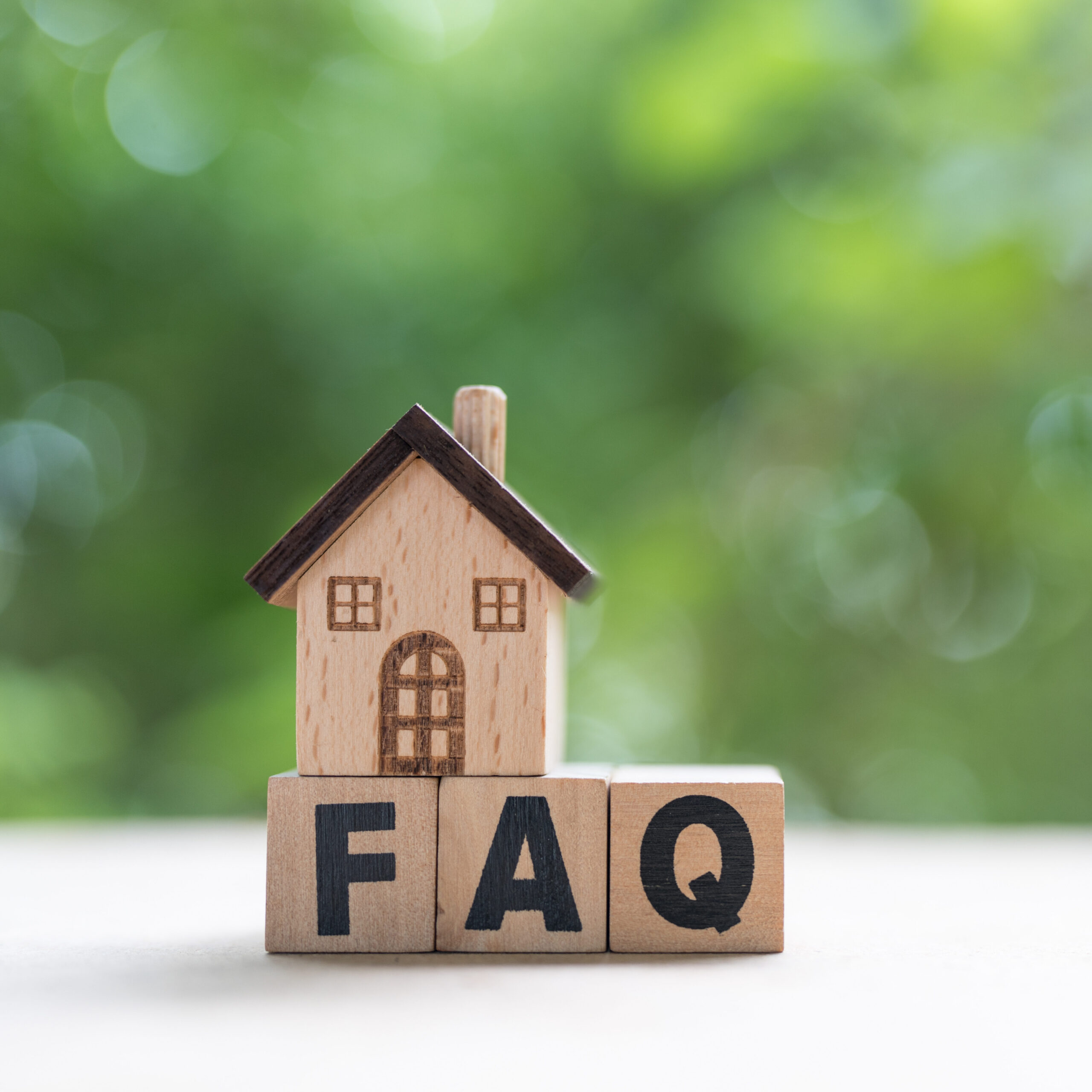 A small wooden house sits on top of three wooden blocks spelling "FAQ," with a blurred green outdoor background.