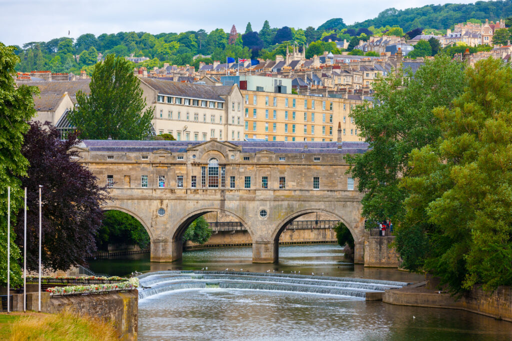 Historic stone bridge with shops spans a river with a curved weir below, surrounded by lush green trees and classic buildings; cityscape and hills visible in the background.