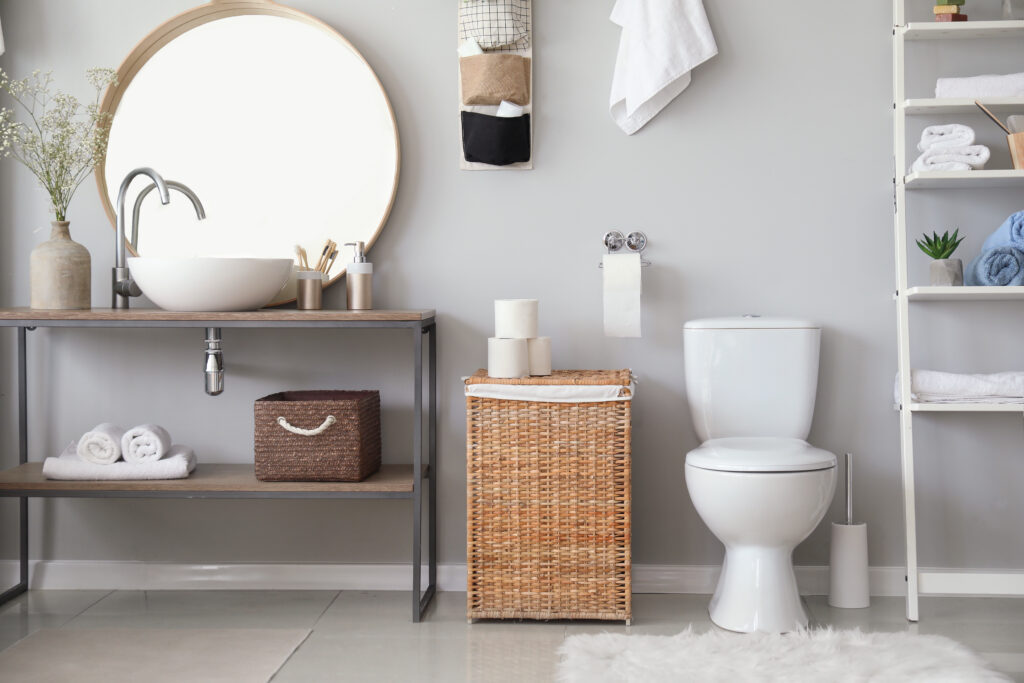 Modern bathroom with a round mirror, vessel sink, wicker basket, white toilet, shelves with towels and toiletries, and light decor against a gray wall.