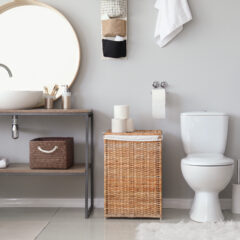 Modern bathroom with a round mirror, vessel sink, wicker basket, white toilet, shelves with towels and toiletries, and light decor against a gray wall.