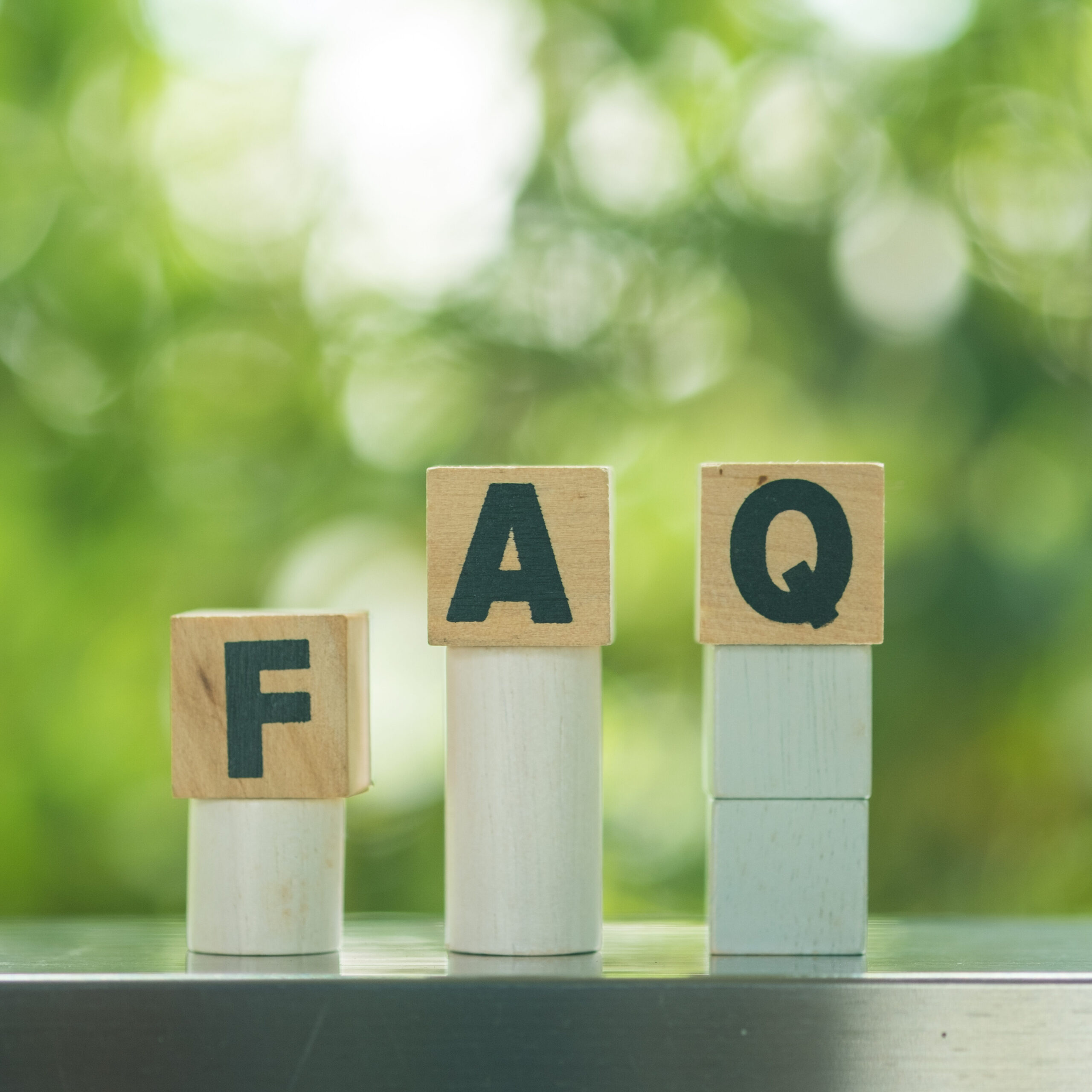 Three wooden blocks with the letters "F," "A," and "Q" are stacked on white and light blue columns, with a blurred green background outdoors.