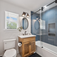 Modern bathroom with a wood vanity, white sink, and black fixtures. There’s a toilet, a window with blinds, and a glass-enclosed shower with blue tiles and a round mirror above the sink.