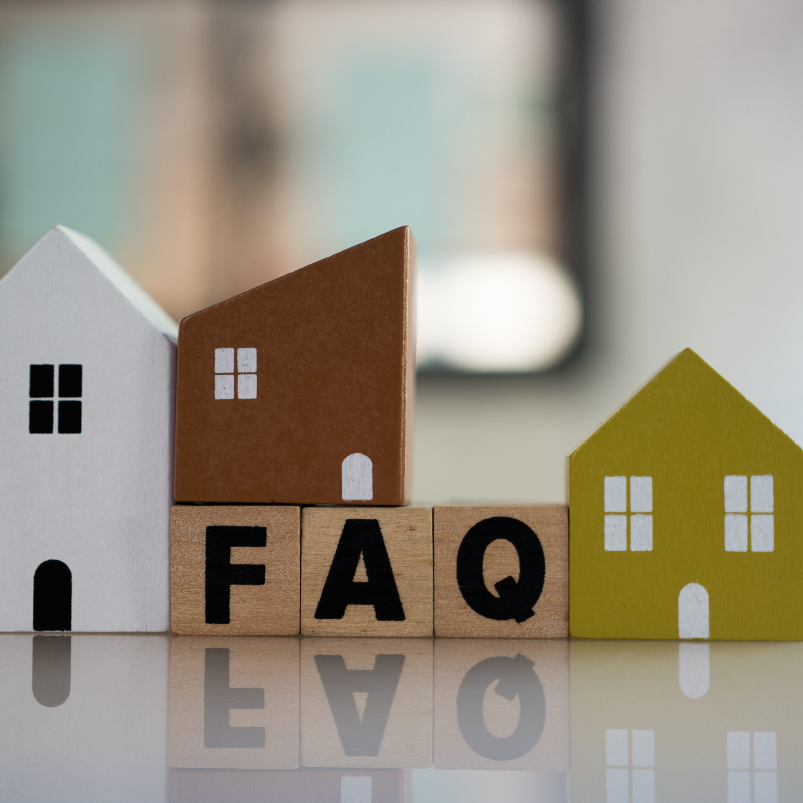 Three wooden house-shaped blocks sit on top of wooden letter blocks spelling "FAQ." The houses are white, brown, and yellow, with small windows and doors, on a reflective surface with a blurred background.