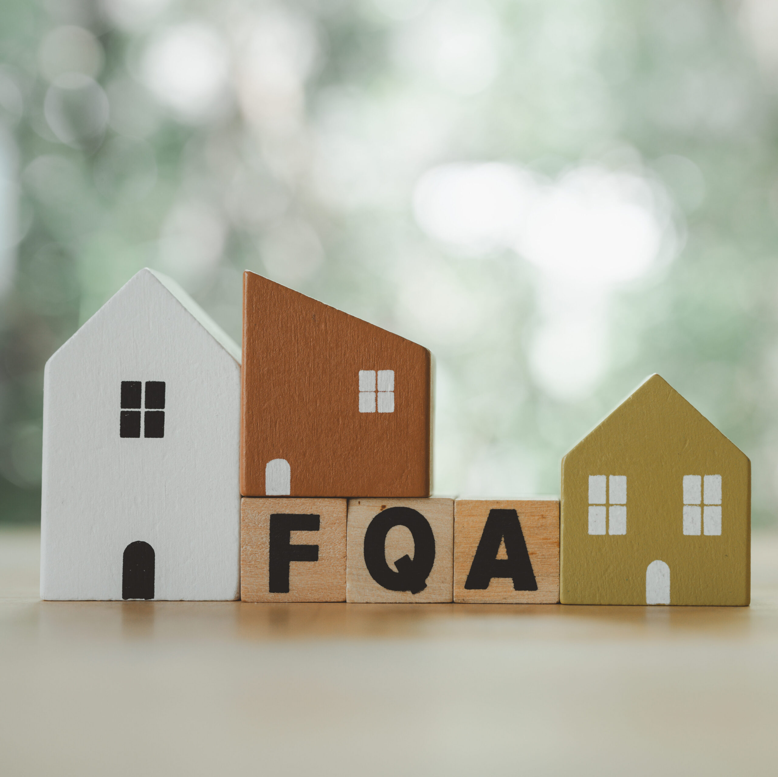 Three small wooden house models in white, brown, and yellow stand behind wooden blocks spelling "FQA," with a blurred green background.